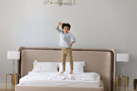 Cheerful little adorable preschool mixed race kid boy jumping on bed, having fun alone iat home. Happy energetic small 6s child son playing in bedroom, enjoying active leisure weekend pastime.の写真素材