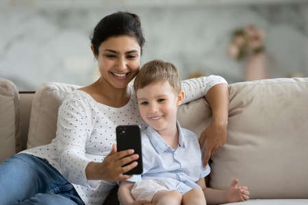 Happy affectionate young asian indian mother and small cute caucasian child son using smartphone, watching funny video in social networks or posing for selfie photo, entertaining online at home.の写真素材