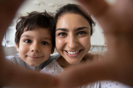 Head shot close up happy bonding young asian indian mother and playful adorable little kid son making heart symbol with fingers, posing for selfie photo or recording funny video, entertaining at home.の写真素材