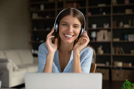 Happy millennial girl wearing headphones, looking at camera over laptop, smiling. Young distance...