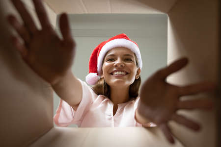 Excited Christmas gift receiver in Santa hat taking present wrap from delivery carton box. Happy smiling girl, young woman outstretching hands inside package. Low angle, bottom view portraitの写真素材
