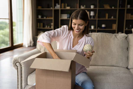 Happy millennial girl unpacking carton box at home, opening package, holding skein of rope, paper container, wrapping, packing parcel. Customer receiving purchase from online store. Delivery serviceの写真素材