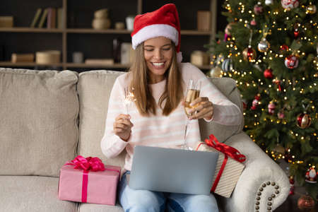 Happy excited young woman in Xmas Santa hat toasting fizzy wine at laptop webcam, holding sparklers. Girl making New Year video call from home, sitting on couch at gifts and festive Christmas treeの写真素材