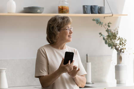 Smiling dreamy middle aged mature woman holding cellphone in hands, spending time in modern kitchen....