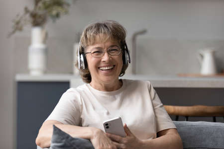 Happy laughing middle aged old woman in eyewear listening f music or affirmations in headphones, choosing tracks in mobile library applications, relaxing on cozy sofa, enjoying hobby activity at home.の写真素材