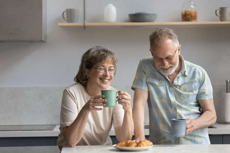 Happy middle aged mature retired married couple drinking hot morning coffee or tea, eating fresh croissants, enjoying communicating during weekend breakfast in modern kitchen, family relations conceptの写真素材