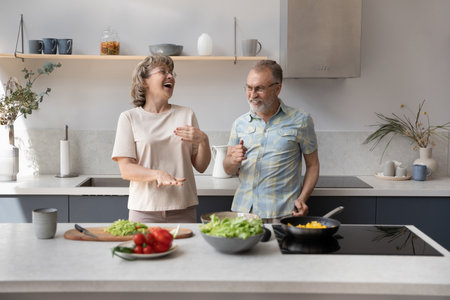 Distracted from preparing healthy food, joyful married middle aged elderly couple dancing to favorite disco music, entertaining together in modern kitchen, having fun enjoying leisure time together.の写真素材