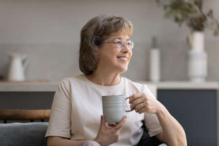 Dreamy middle aged old woman in eyeglasses holding cup of tea or morning coffee sitting on...