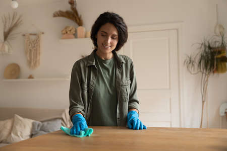 Professional cleaner, housewife, maid cleaning furniture in apartment. Happy Hispanic woman in rubber gloves washing tabletop surface in living room, smiling, enjoying housework. Household conceptの写真素材