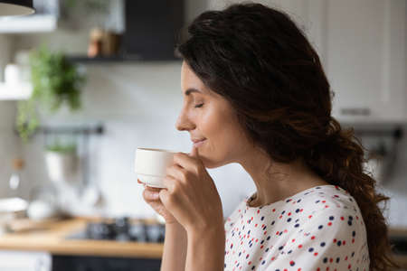 Happy peaceful woman enjoying morning coffee in kitchen, holding cup, inhaling aroma with closed eyes. Homeowner relaxing over cup of hot beverage at home, smiling, thinking and dreamingの写真素材
