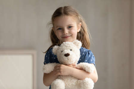 Portrait of smiling adorable preschool small kid girl cuddling favorite fluffy toy, posing alone at home. Happy cute 7s little child daughter holding teddy bear, feeling cheerful in kindergarten.の写真素材
