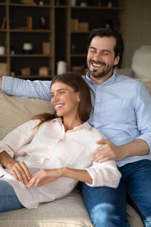 Vertical image happy laughing sincere loving affectionate married couple resting together on cozy couch, enjoying carefree leisure weekend time together in own house, good family relations concept.の写真素材