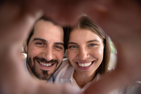 Close up head shot smiling sincere young family couple making heart symbol with fingers to camera, showing affectionate feelings, devotion and care, candid relationship, true love, donation concept.の写真素材