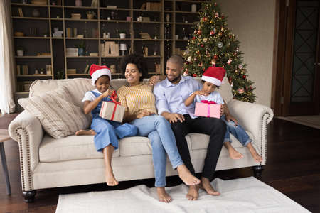 Happy loving young african american couple parents watching small kids siblings unpacking wrapped gifts, sitting together on cozy sofa near decorated tree, New Year or Merry Christmas celebration.の写真素材