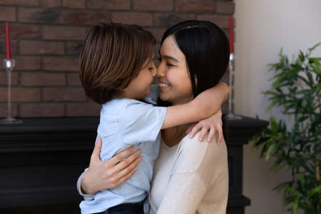 Happy Asian mixed race mom holding son boy in arms, hugging kid with noses touch, smiling, enjoying warm moment. Younger mum carrying kid and dancing at home. Motherhood, adoption, family conceptの写真素材