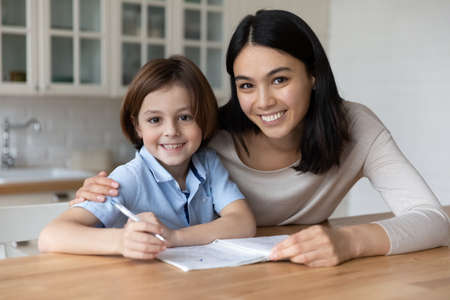 Head shot portrait of happy mom and schoolboy son doing school homework together. Young mother giving support to kid studying at home, taking notes in copybook, learning to write. Parenthood conceptの写真素材
