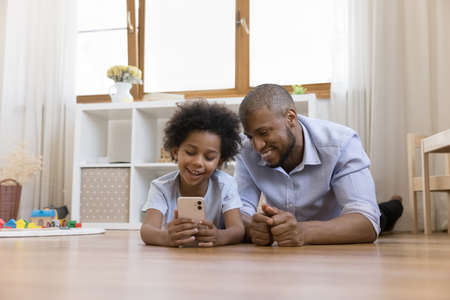 Happy small adorable african american child boy showing funny mobile application or playing games to caring young father, relaxing lying together on warm heated wooden floor at home, tech addiction.の写真素材