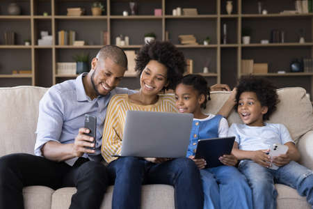 Happy young african american couple parents and cute little children using different gadgets, spending time online, playing games, entertaining sitting together on sofa in living room, tech addiction.の写真素材
