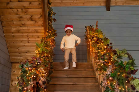 Smiling small African American boy child run down beautiful decorated stairs feel excited on Christmas Eve at home. Happy little biracial kid have fun enjoy New Year winter holidays in cozy house.の写真素材