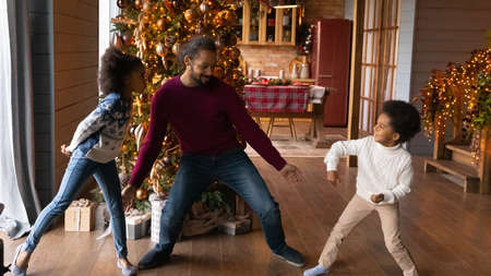 Wide banner panoramic view of overjoyed young African American father and two small children dancing together near Christmas tree at home. Happy biracial dad and kids have fun on winter holidays.の写真素材