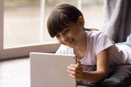 Happy gen Z kid girl watching movie on tablet computer, resting on heating floor at window, reading...
