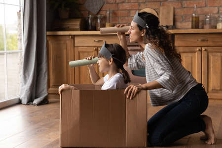 Happy creative mother and active daughter girl playing pirates ship at home, sailing carton box boat, looking forward through toy paper spyglasses, enjoying home activities. Family role gameの写真素材
