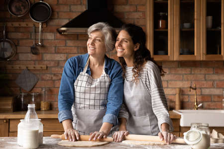 Happy mature 60s mother and grown daughter woman baking pie, cookies family lunch, dinner in kitchen, rolling dough on table, looking away, thinking, smiling. Family, intergenerational friendshipの写真素材
