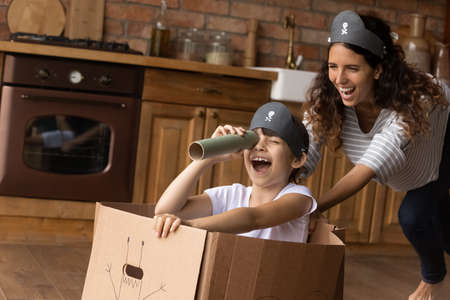 Excited kid and happy laughing mom playing pirates at home, sailing toy paper ship, looking forward through spyglasses, dragging carton box sailboat on floor, laughing, enjoying role game, amusementの写真素材