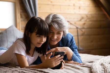 Happy generation Z girl and mature senior 60s grandma using smartphone together, making video call, talking to family, smiling at screen. Granddaughter teaching grandma to use online virtual appの写真素材