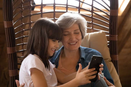 Smiling gen Z granddaughter girl teaching senior grandma to use online app on smartphone. Granny and grandkid sitting together in armchair, holding mobile phone, making video call, watching mediaの写真素材