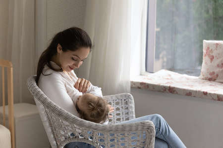 Loving young mom breastfeeding little daughter, sitting in cozy chair at home, happy caring mother...