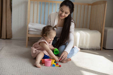 Happy young mother and little daughter playing with toys, sitting on warm floor in nursery at home...