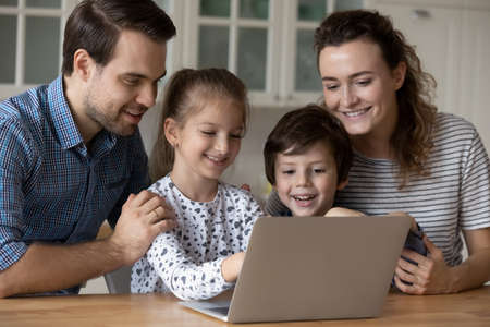 Happy two small kids boy girl choosing goods in internet store with joyful couple parents, using educational computer applications, plying games together or watching cartoons online in modern kitchen.の写真素材