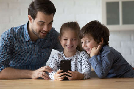 Happy millennial father using funny cellphone applications with adorable small children siblings,...