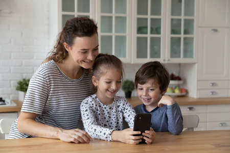 Happy caring millennial mother controlling small children son daughter using cellphone applications, playing online mobile video games, watching funny photo video content in social network in kitchen.の写真素材