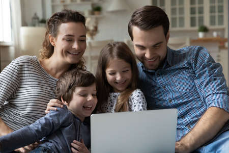 Laughing affectionate two generations family couple parents and little son and daughter using computer, choosing goods shopping online, web surfing, watching cartoons, relaxing on sofa at home.の写真素材