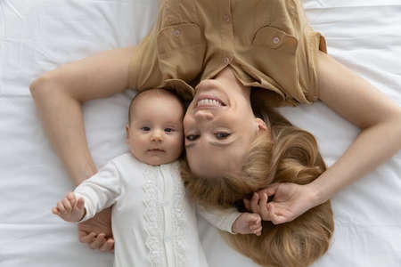 Top view head shot portrait of smiling young mother and baby lying on comfortable bed, white sheet, happy mom and newborn child infant looking at camera, posing for family portrait at homeの写真素材