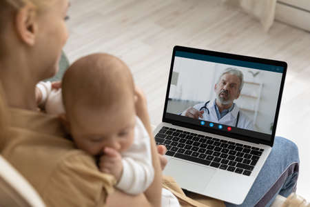 Caring young mother holding sleeping baby, using laptop, making video call to mature man doctor at...