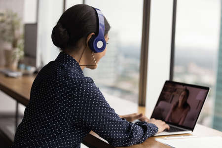 Concentrated young indian businesswoman in headphones holding video call conversation with female colleague on computer, discussing working issues. Skilled hr manager interviewing candidate distantly.の写真素材