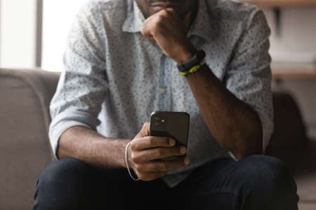 Cropped image thoughtful young african mixed race man using cellphone, feeling anxious getting spam scam message or email with bad news, dissatisfied with wrong device work, tech addiction concept.の写真素材