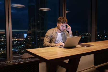 Thoughtful young man working online on computer, sitting at table late in night in modern office room, analyzing electronic documents, considering problem solution, communicating with clients.の写真素材