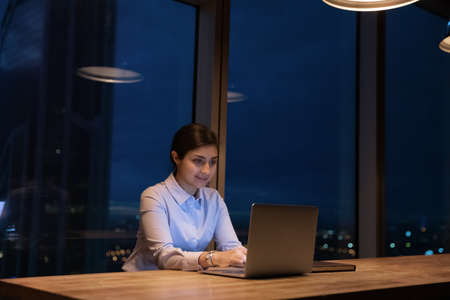 Smiling beautiful young female indian manager employee working on computer, creating new online project web surfing or communicating distantly late in evening in modern office with panoramic window.の写真素材