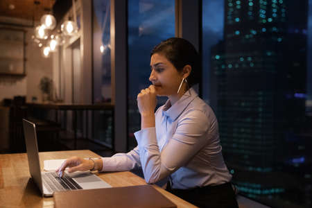 Concentrated young indian businesswoman brainstorming project ideas or considering problem solution, working overtime on laptop late in evening alone in modern office with panoramic cityscape view.の写真素材