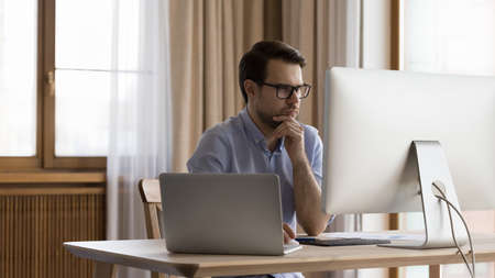 Concentrated young 30s ceo manager businessman in eyewear looking at computer monitor, considering...