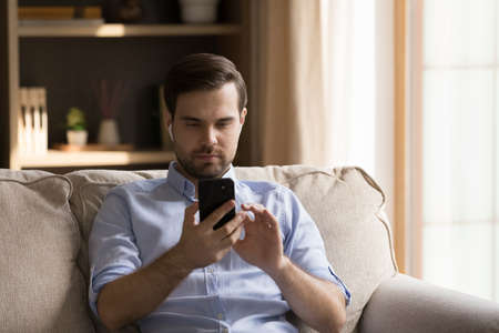 Happy young handsome man in wireless earbuds choosing music in mobile playlist, listening audio tracks, watching video in social network or holding web camera call conversation using cellphone at homeの写真素材