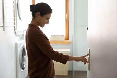 Young Indian ethnicity female push button turns off on light inside modern domestic laundry room,...
