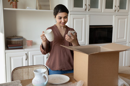 Indian woman standing in kitchen unpacks parcel box with new crockery items, holding coffee cup and...