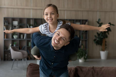 Loving father piggy backing adorable 8s daughter pretending flying with arms outstretched, looking in distance, dreaming, smiling dad with little girl child engaged in funny activity, having funの写真素材