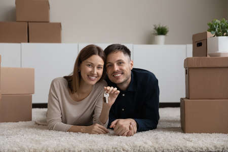Portrait of happy married couple showing demonstrating keys and looking at camera, excited by relocation, lying on floor in new home, smiling woman and man celebrating moving day, relocationの写真素材