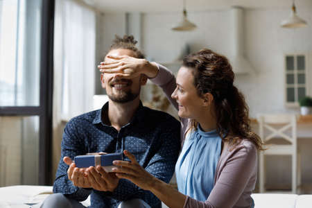 Happy young loving woman covering eyes of curious sincere husband, making surprise preparing wrapped present for birthday or wedding anniversary, congratulating with special occasion at home.の写真素材